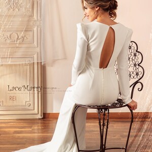 May include: A white long-sleeved wedding dress with a low back and a train. The dress is fitted and has a simple design. The model is wearing the dress and sitting on a chair.