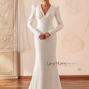 May include: A white, long-sleeved, v-neck wedding dress with a fitted bodice and a flowing skirt. The dress has a simple, elegant design with a slight puff at the shoulders.