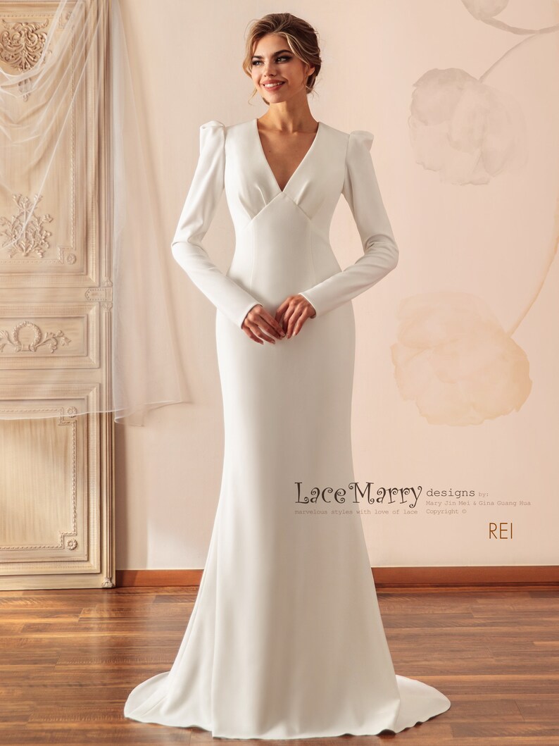 May include: A white, long-sleeved, v-neck wedding dress with a fitted bodice and a flowing skirt. The dress has a simple, elegant design with a slight puff at the shoulders.