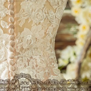 May include: A close-up of a white lace wedding dress with a row of buttons down the back. The dress is made of delicate lace with a floral pattern. The dress is designed by Mary Jin Mel & Gina Guang Hua.