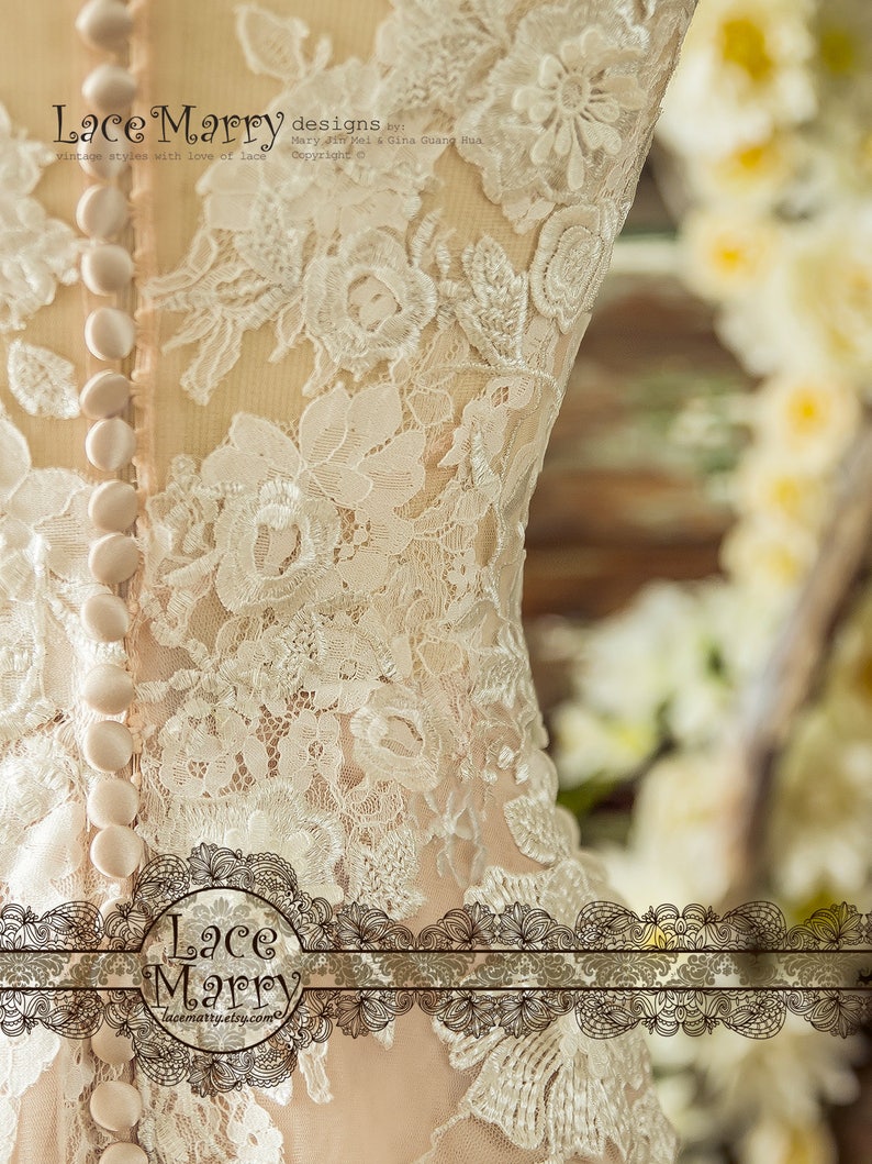 May include: A close-up of a white lace wedding dress with a row of buttons down the back. The dress is made of delicate lace with a floral pattern. The dress is designed by Mary Jin Mel & Gina Guang Hua.