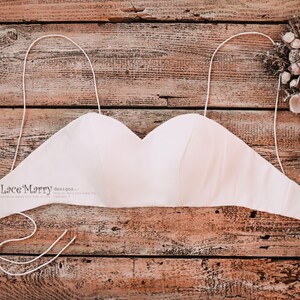 Pu&ograve; includere: Un top da sposa bianco senza spalline con spalline regolabili. Il top &egrave; realizzato in un tessuto morbido e setoso e ha una scollatura a cuore. Il top &egrave; appoggiato su una superficie di legno.