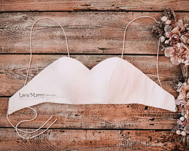 Pu&ograve; includere: Un top da sposa bianco senza spalline con spalline regolabili. Il top &egrave; realizzato in un tessuto morbido e setoso e ha una scollatura a cuore. Il top &egrave; appoggiato su una superficie di legno.