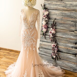 May include: A blush pink wedding dress with long sleeves and intricate white lace detailing. The dress has a fitted bodice and a flowing skirt.