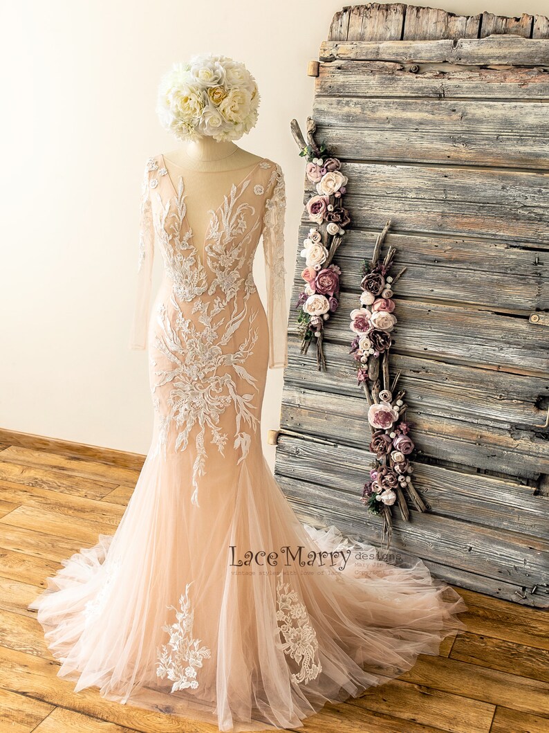 May include: A blush pink wedding dress with long sleeves and intricate white lace detailing. The dress has a fitted bodice and a flowing skirt.