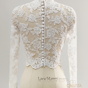 May include: A white lace bridal top with long sleeves and a high neckline. The back features a row of small, white buttons. The intricate floral lace design is visible against a sheer fabric. The top is displayed on a mannequin.