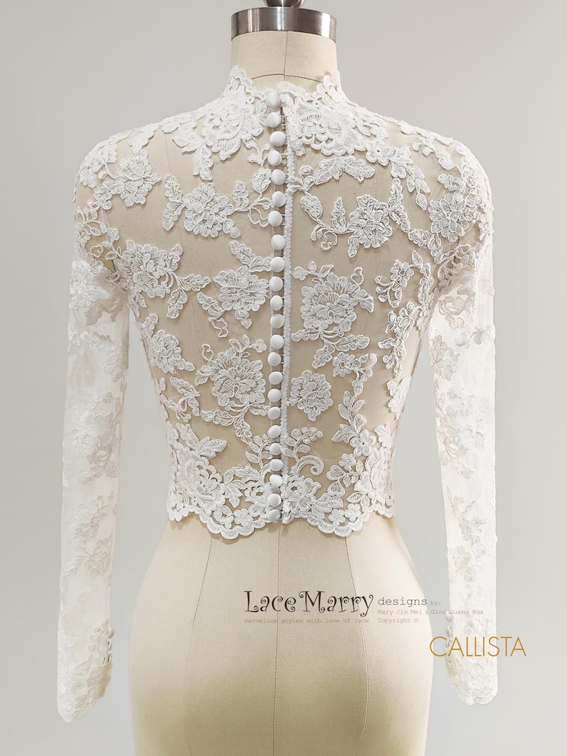 May include: A white lace bridal top with long sleeves and a high neckline. The back features a row of small, white buttons. The intricate floral lace design is visible against a sheer fabric. The top is displayed on a mannequin.