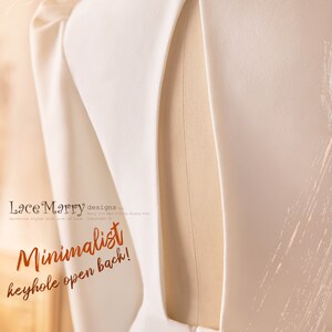 May include: A white wedding dress with a keyhole open back. The dress is on a white dress form. The text "Minimalist keyhole open back!" is superimposed on the image.