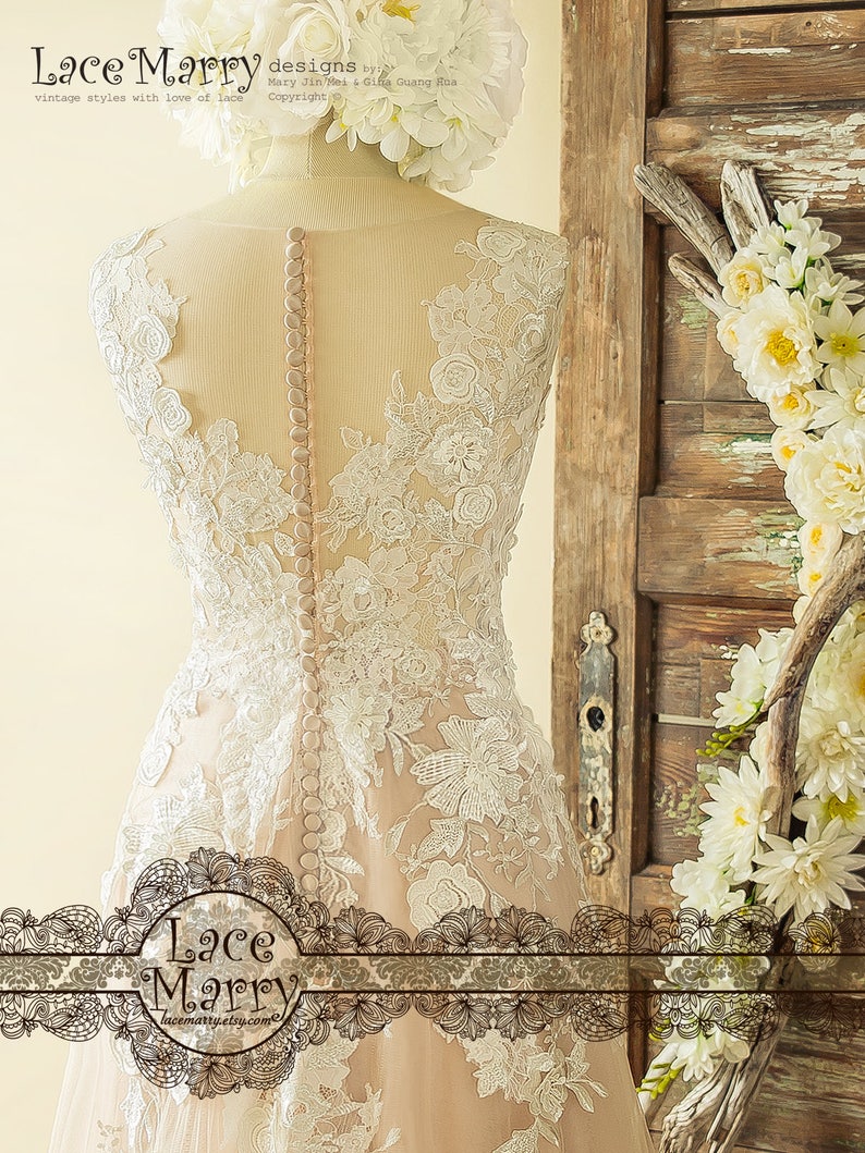 May include: A white lace wedding dress with a nude lining and a row of buttons down the back. The dress has a fitted bodice and a flowing skirt. The dress is displayed on a mannequin against a wooden background.