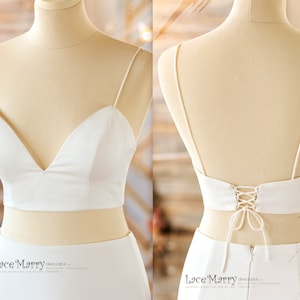 May include: A white bridal crop top with a deep V-neckline and spaghetti straps. The back features a lace-up design. The top is paired with a matching white skirt, creating a modern wedding ensemble. The fabric appears smooth and elegant.