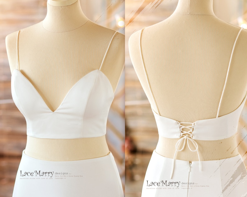 May include: A white bridal crop top with a deep V-neckline and spaghetti straps. The back features a lace-up design. The top is paired with a matching white skirt, creating a modern wedding ensemble. The fabric appears smooth and elegant.