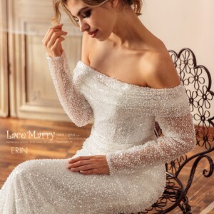 May include: A white off-the-shoulder wedding dress with long sleeves and a beaded design. The dress is fitted and has a long train. The model is sitting on a black metal chair. The text "Lace Marry designs by Mary Jin Mei & Gina Gu marvelous styles with love of Lace Copyright &copy; ERIN" is at the bottom of the image.