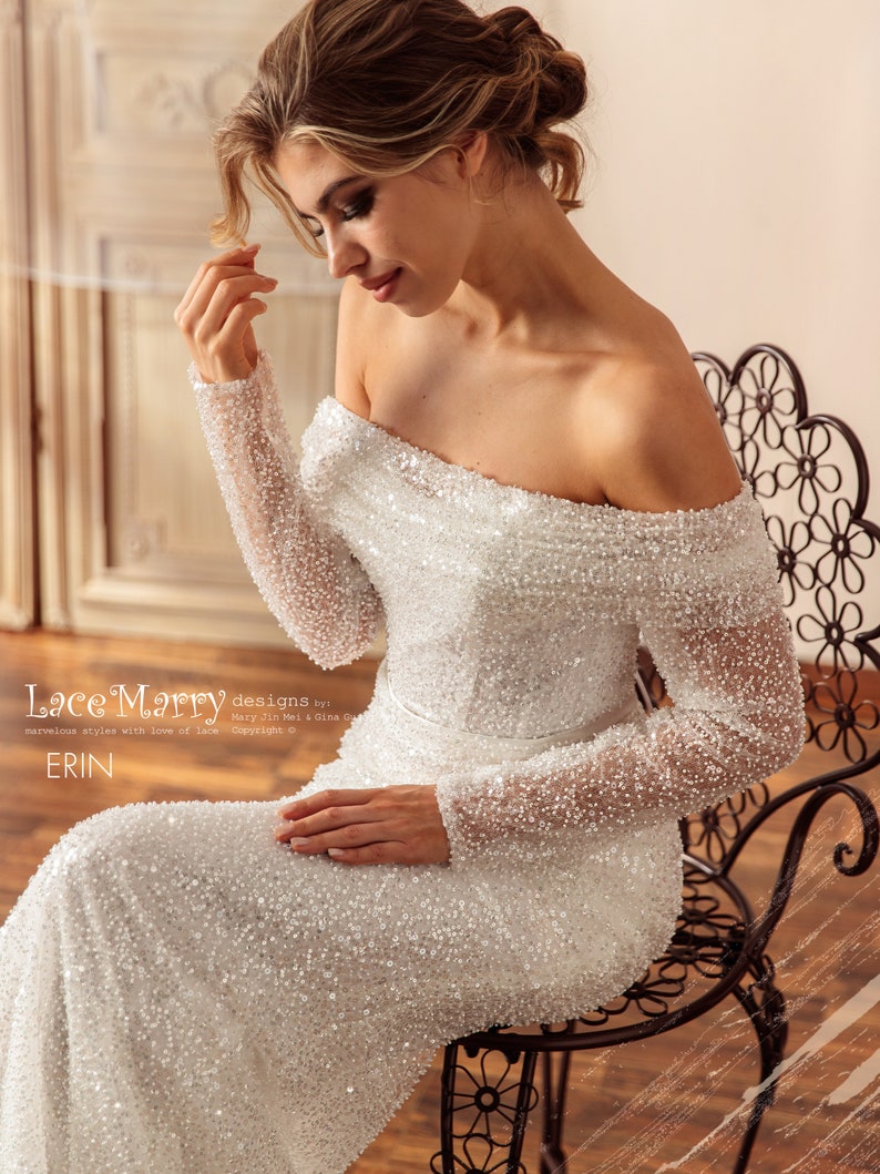 May include: A white off-the-shoulder wedding dress with long sleeves and a beaded design. The dress is fitted and has a long train. The model is sitting on a black metal chair. The text "Lace Marry designs by Mary Jin Mei & Gina Gu marvelous styles with love of Lace Copyright &copy; ERIN" is at the bottom of the image.