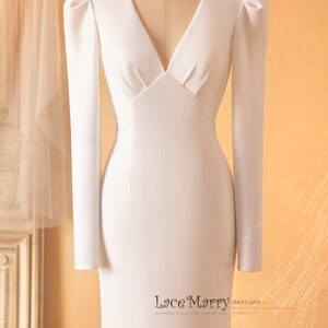 May include: A white, long-sleeved, v-neck wedding dress with a fitted bodice and a flowing skirt. The dress has puffed sleeves and a fitted waist.