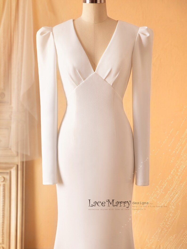 May include: A white, long-sleeved, v-neck wedding dress with a fitted bodice and a flowing skirt. The dress has puffed sleeves and a fitted waist.