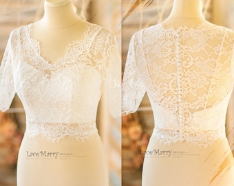 May include: A white lace bridal top with a V-neckline and short sleeves. The back of the top features a row of buttons. The delicate lace design is visible on the front and back of the top. The top is displayed on a mannequin.