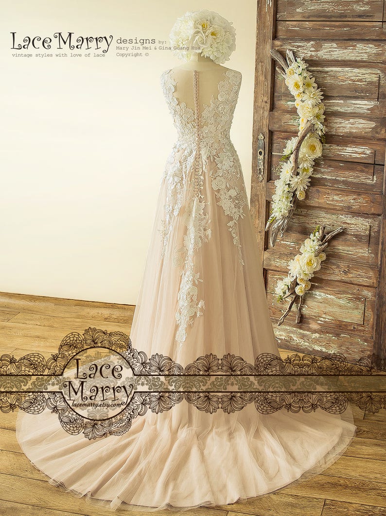 May include: A blush pink tulle wedding dress with white lace appliques and a lace-up back. The dress has a fitted bodice and a flowing skirt. The dress is photographed on a mannequin in front of a wooden door with floral decorations.