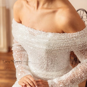 May include: A white off-the-shoulder wedding dress with long sleeves, covered in sequins. The dress has a fitted bodice and a flowing skirt. The dress is designed by Lace Marry designs by Erin.