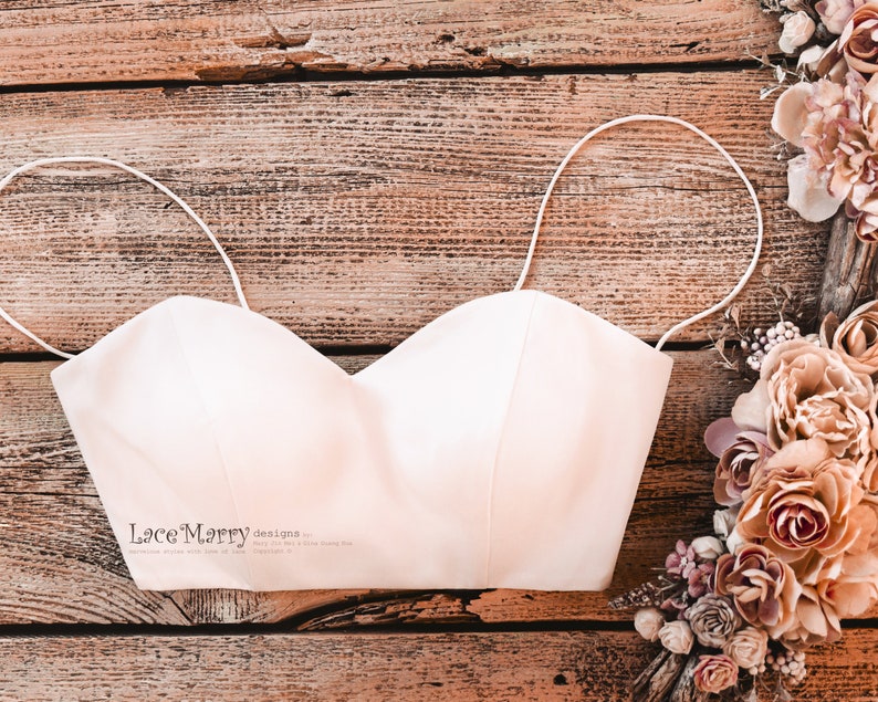 Pu&ograve; includere: Un top bianco senza spalline con spalline sottili, appoggiato su una superficie di legno. Il top &egrave; realizzato in un tessuto liscio e satinato. Il testo "Lace Marry Designs" &egrave; stampato sul davanti del top.
