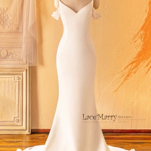 May include: A white wedding dress with a fitted bodice and a flowing skirt. The dress has a sweetheart neckline and off-the-shoulder straps with lace detailing. The dress is displayed on a mannequin against a light brown background.