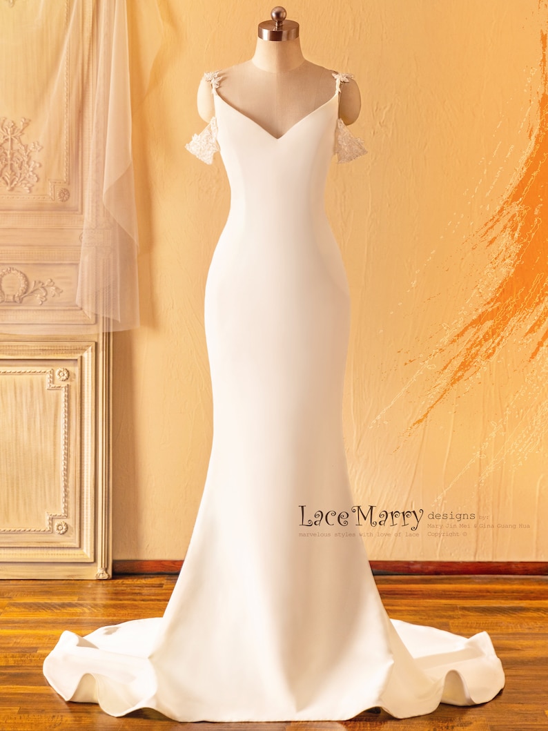 May include: A white wedding dress with a fitted bodice and a flowing skirt. The dress has a sweetheart neckline and off-the-shoulder straps with lace detailing. The dress is displayed on a mannequin against a light brown background.