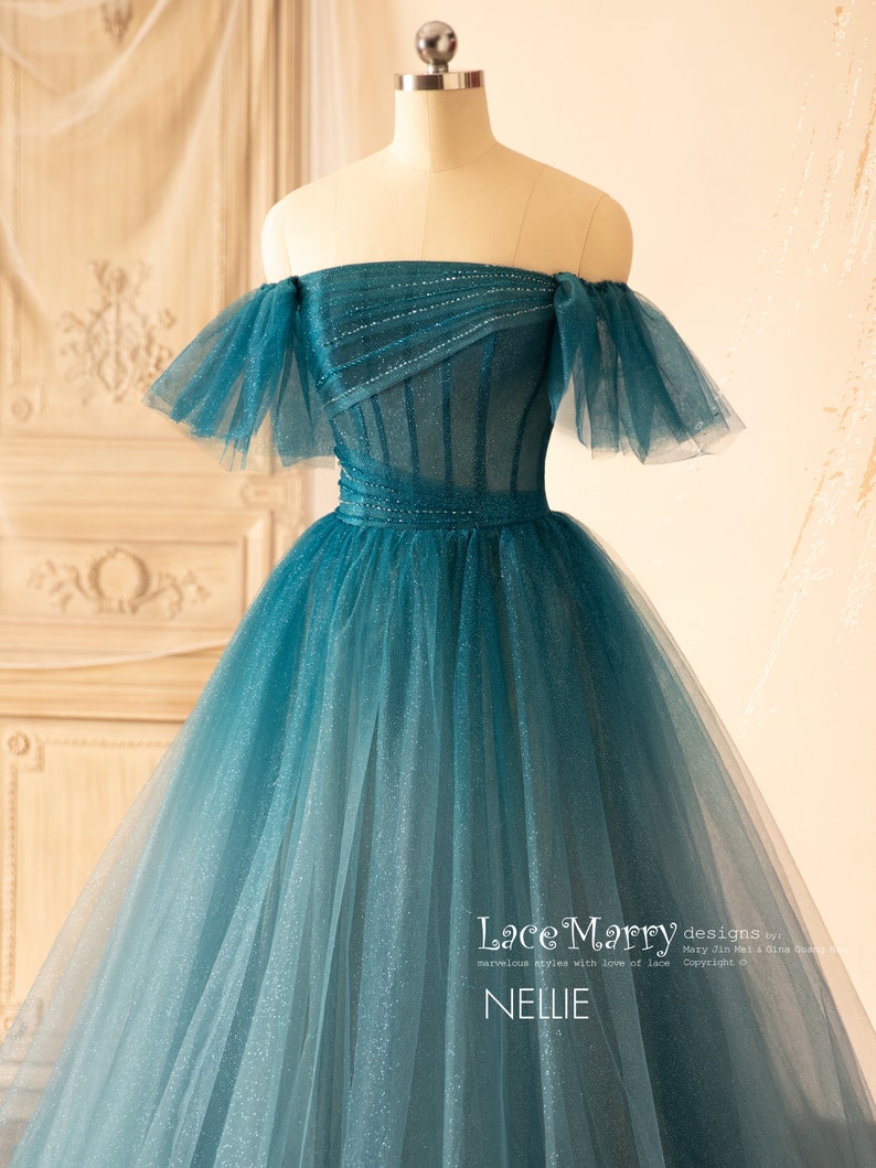 K&ouml;nnte beinhalten: Ein blaues T&uuml;ll-Ballkleid mit offener Schulter, einem figurbetonten Oberteil und einem vollem Rock. Das Oberteil ist mit Pailletten verziert und der Rock hat einen Farbverlauf, der von einem dunkleren Blau an der Oberseite zu einem helleren Blau an der Unterseite &uuml;bergeht. Das Kleid wurde von Lace Marry entworfen.