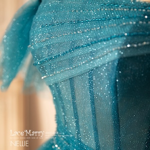 K&ouml;nnte beinhalten: Ein t&uuml;rkisfarbenes T&uuml;llkleid mit einem tr&auml;gerlosen Oberteil und einem glitzernden, perlenbesetzten Design. Das Kleid hat ein figurbetontes Oberteil und einen flie&szlig;enden Rock.