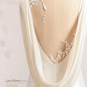 Pu&ograve; includere: Un abito da sposa in satin bianco con schiena scoperta e un pannello trasparente con delicati ricami di pizzo floreale. L'abito ha una schiena drappeggiata a scialle.