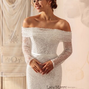May include: A white off-the-shoulder wedding dress with long sleeves and a fitted silhouette. The dress is covered in small sequins and has a satin sash at the waist. The dress is designed by Lace Marry.
