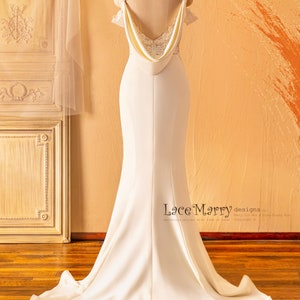 May include: A white wedding dress with a lace bodice and a flowing skirt. The dress has a low back with a draped neckline and a fitted silhouette. The dress is shown on a mannequin against a light brown background.