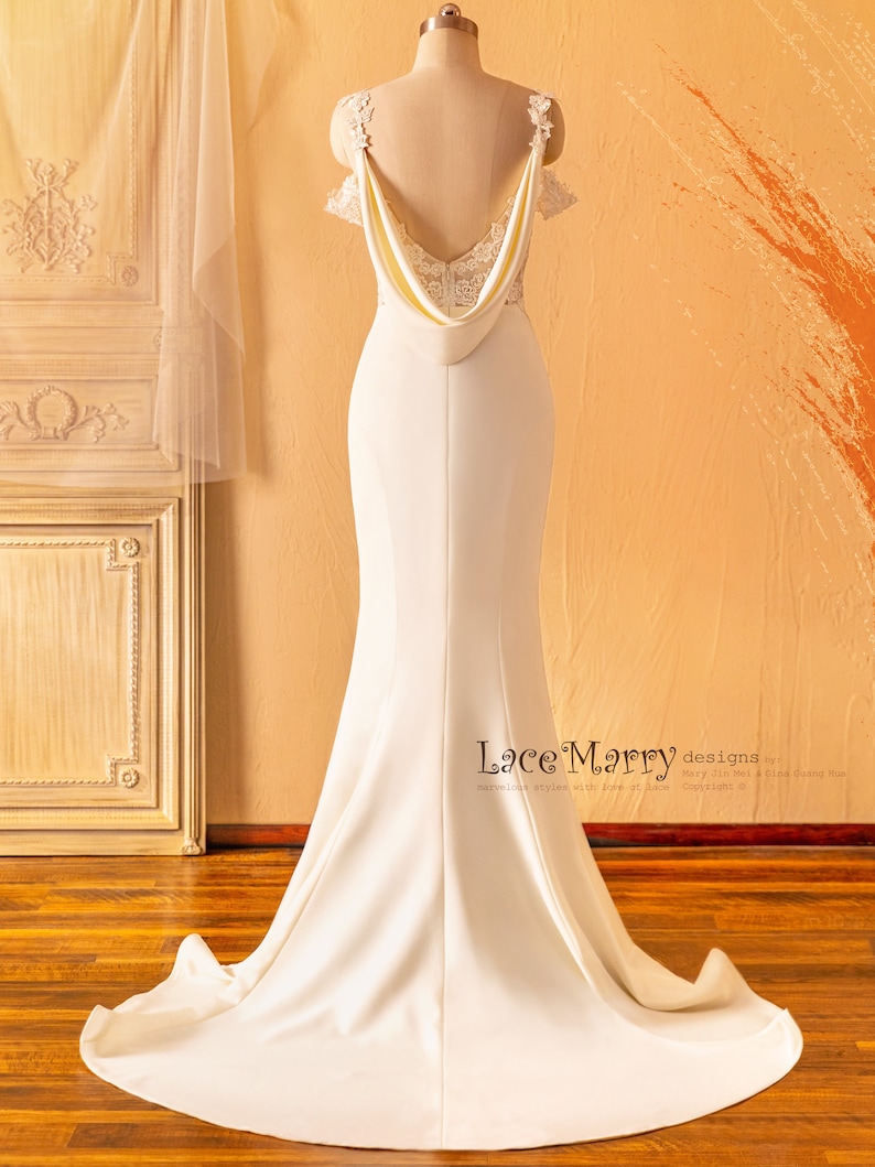 May include: A white wedding dress with a lace bodice and a flowing skirt. The dress has a low back with a draped neckline and a fitted silhouette. The dress is shown on a mannequin against a light brown background.