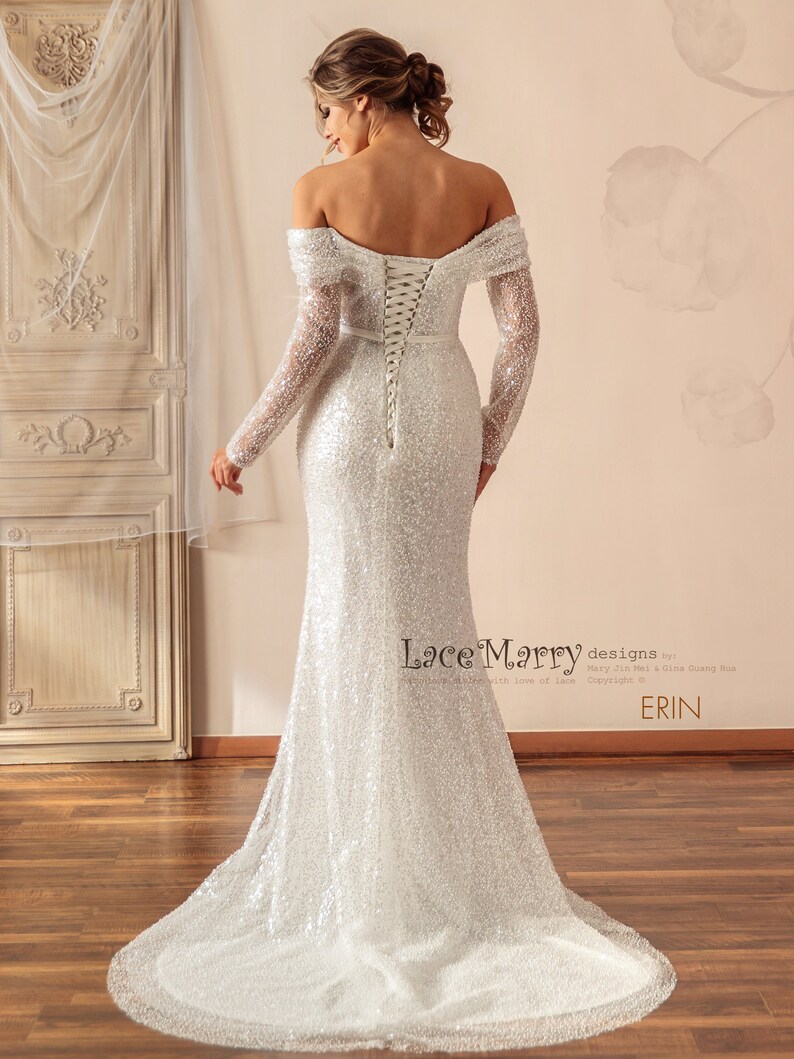 May include: A white off-the-shoulder sequined wedding dress with long sleeves and a lace-up back. The dress has a fitted bodice and a flowing skirt. The dress is designed by Lace Marry.
