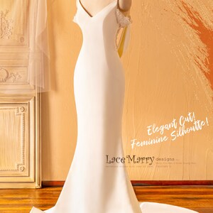 May include: A white wedding dress with a fitted bodice and a flowing skirt. The dress has a V-neckline and delicate lace straps. The dress is on a mannequin and the background is a light brown wall.