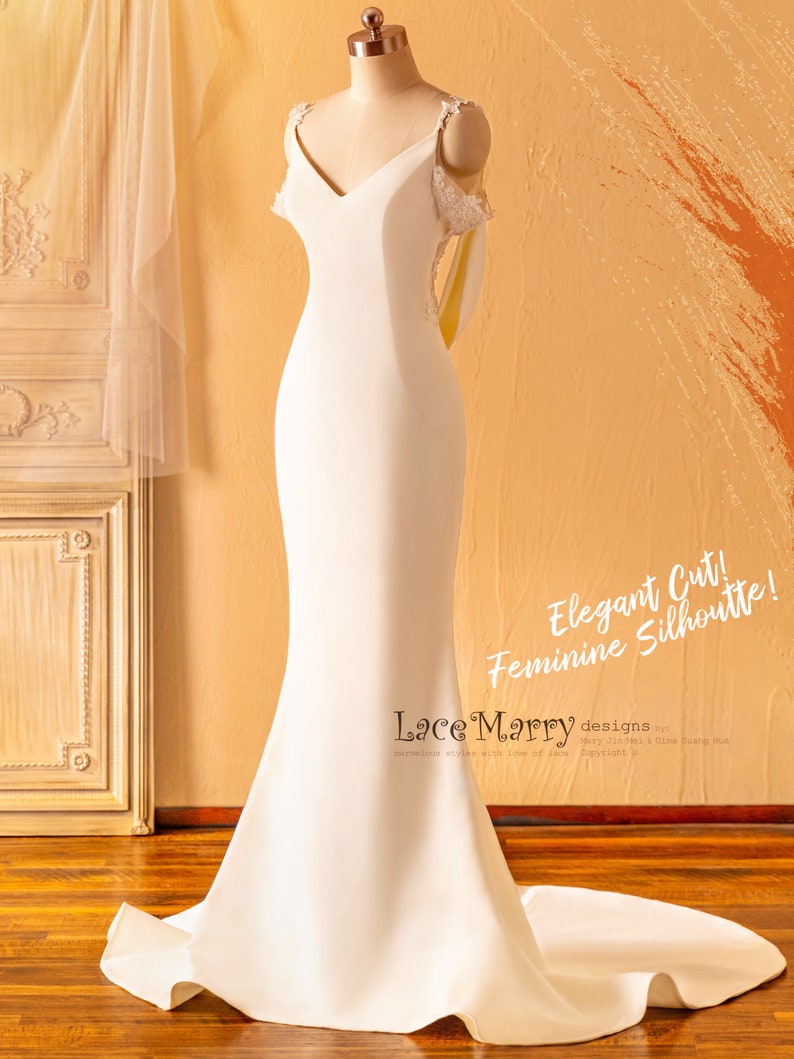 May include: A white wedding dress with a fitted bodice and a flowing skirt. The dress has a V-neckline and delicate lace straps. The dress is on a mannequin and the background is a light brown wall.