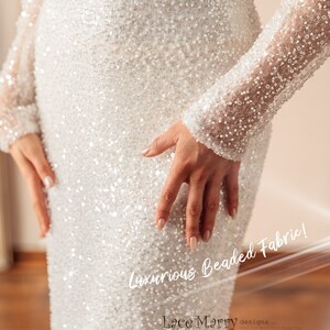 May include: A white beaded gown with long sleeves. The gown is covered in small, white beads that create a shimmering effect. The text "Luxurious Beaded Fabric!" is written on the gown. The text "Lace Marry designs" is written on the bottom of the gown.
