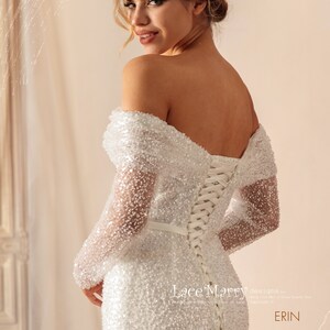 May include: A white off-the-shoulder wedding dress with long sleeves and a lace-up back. The dress is covered in small sequins and has a fitted silhouette. The dress is labeled "Lace Mary designs by... Erin".
