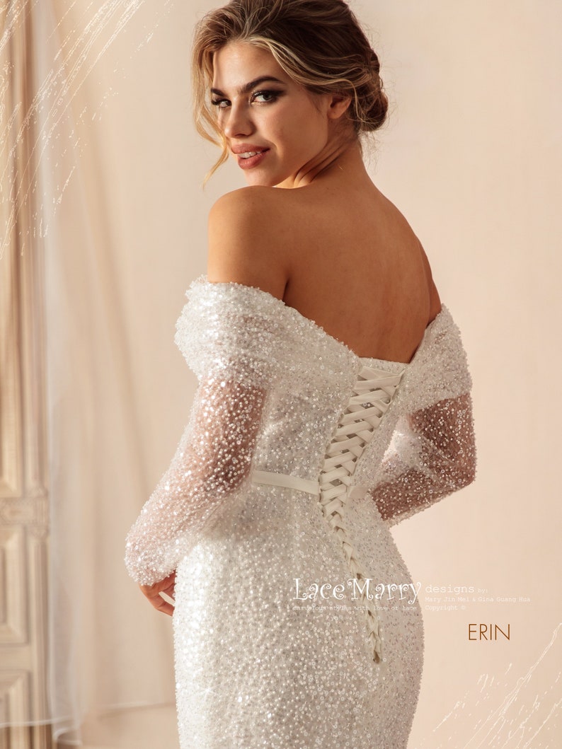 May include: A white off-the-shoulder wedding dress with long sleeves and a lace-up back. The dress is covered in small sequins and has a fitted silhouette. The dress is labeled "Lace Mary designs by... Erin".