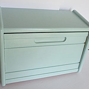 May include: A light sage green wooden bread box with a smooth matte finish. The rectangular container features a hinged lid with a horizontal recessed handle across the front panel and a clean, simple design with rounded side edges.