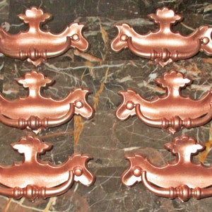 May include: Six antique-style cabinet pulls in a copper finish. Each pull features a decorative, ornate design with a curved handle.