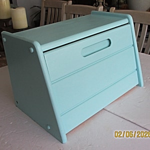 May include: A light blue wooden bread box with a sloped lid and a handle. The box has three horizontal slats on the front and is painted in a solid, matte finish. The box is sitting on a white surface.