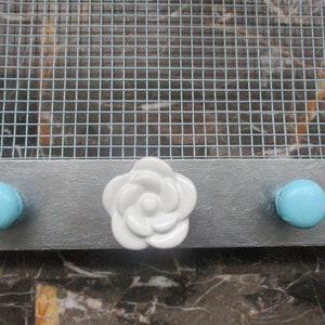 May include: A decorative wall-mounted organizer with a silver-painted wooden base. It features two light blue knobs and a white ceramic flower-shaped knob. A metal mesh grid is attached above the base.