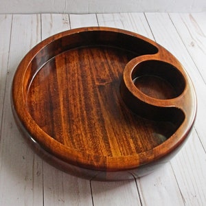 May include: A round wooden serving tray with a smaller round bowl carved into the side. The tray is dark brown and has a glossy finish.