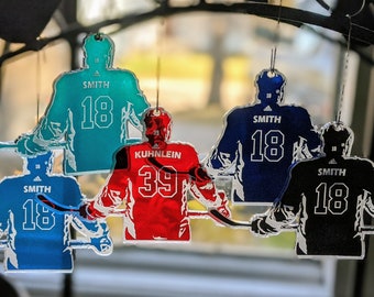 May include: Five hockey player ornaments, each with a different color and player name. The ornaments are made of clear acrylic and feature a silhouette of a hockey player in a skating position. The player names are "Smith 18", "Smith 18", "Smith 18", "Smith 18", and "Kuhlein 39".