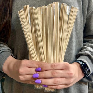 May include: A collection of light-coloured wooden diffuser sticks, packaged in clear plastic. The sticks are held in front of a person wearing a grey sweatshirt. The person's nails are painted a bright purple colour.