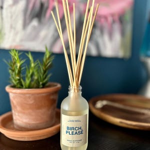 May include: A clear glass reed diffuser bottle with the text "BIRCH, PLEASE" on a white label. The bottle contains a light-colored liquid and several wooden reeds. A small potted plant and a wooden tray are also visible.