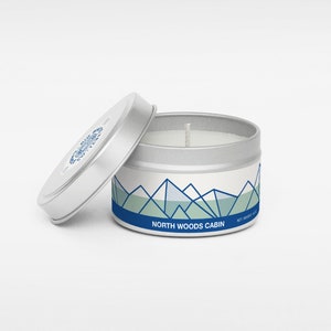 Pu&ograve; includere: Una candela in una lattina argentata con un'etichetta bianca che presenta un design di montagna blu e verde. L'etichetta recita "North Woods Cabin".
