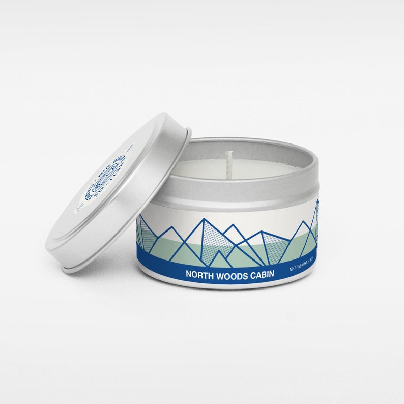 Pu&ograve; includere: Una candela in una lattina argentata con un'etichetta bianca che presenta un design di montagna blu e verde. L'etichetta recita "North Woods Cabin".