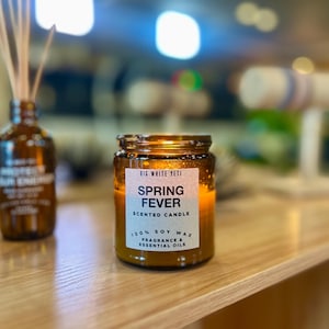 May include: A lit, amber-colored scented candle in a glass jar. The label reads "Spring Fever" and "100% Soy Wax." The candle sits on a light-colored wooden surface, with a blurred background.