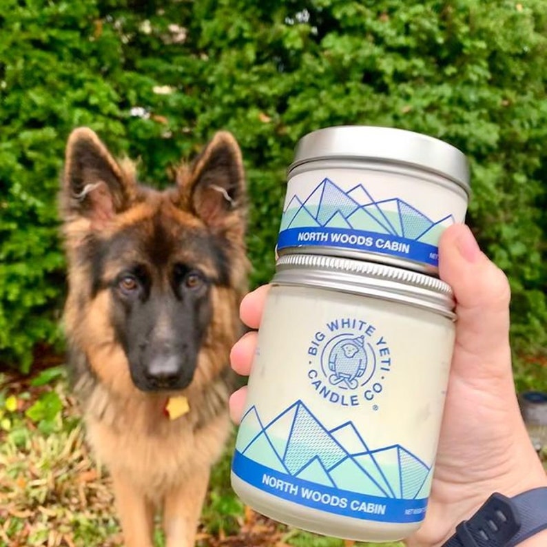 Pu&ograve; includere: Un barattolo di candela bianco con un design di montagna blu e verde e il testo "Big White Yeti Candle Co." e "North Woods Cabin".