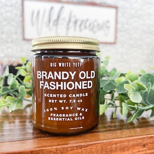 May include: A brown glass candle jar with a gold lid. The label reads "BIG WHITE YETI BRANDY OLD FASHIONED SCENTED CANDLE NET WT. 7.5 oz 100% SOY WAX FRAGRANCE & ESSENTIAL OILS".